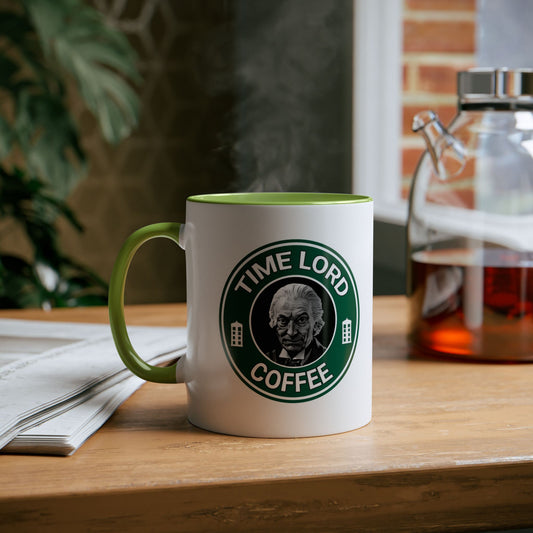 A white ceramic coffee mug with a green handle and interior, featuring a circular green and white 'TIME LORD COFFEE' logo on the front. The mug is shown on a wooden table with a glass carafe of tea in the background.